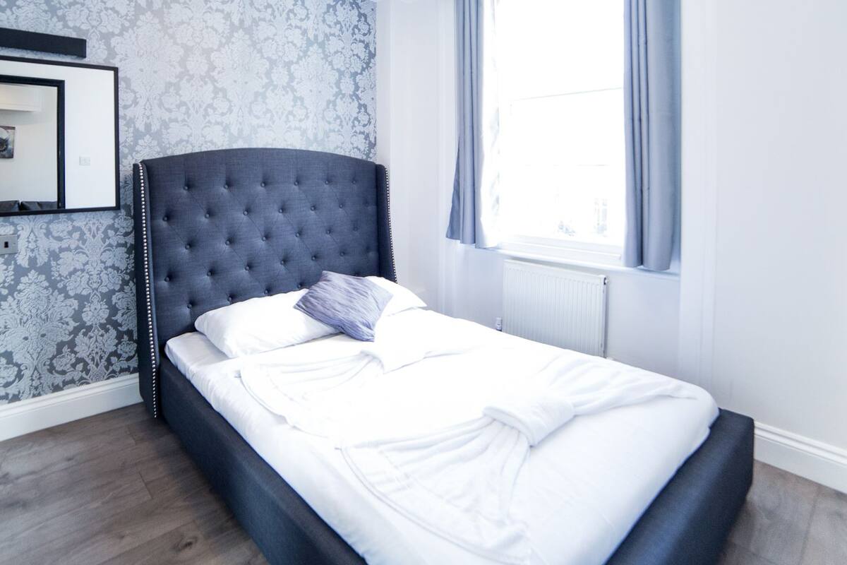 A neatly made bed with white linens and a grey headboard in a room with patterned wallpaper and a window with curtains.