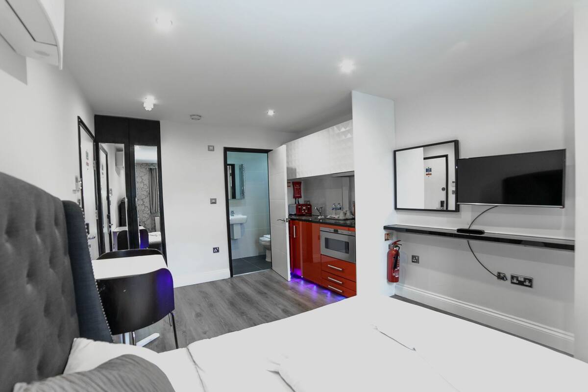 A modern hotel room with a bed, a desk, a chair, a TV, a kitchen area, and a bathroom.