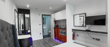 A modern hotel room with a bed, a desk, a chair, a TV, a kitchen area, and a bathroom.