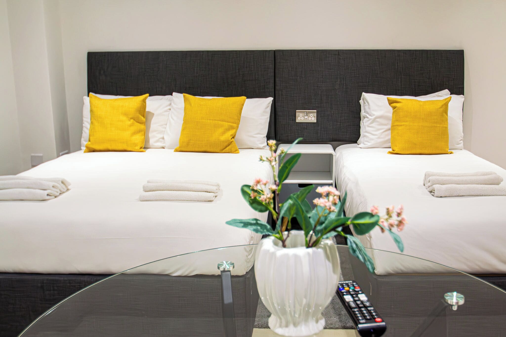 A hotel room with two beds, each with white bedding and yellow pillows, a glass coffee table with a vase of flowers, and a remote control on the table.
