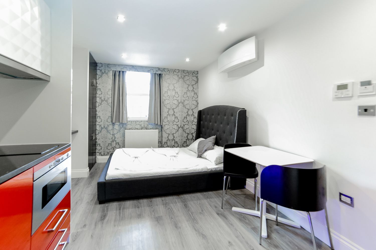 Un dormitorio moderno con cama, mesita de noche y escritorio. Se observa una ventana con cortinas, un radiador y un aire acondicionado de pared.