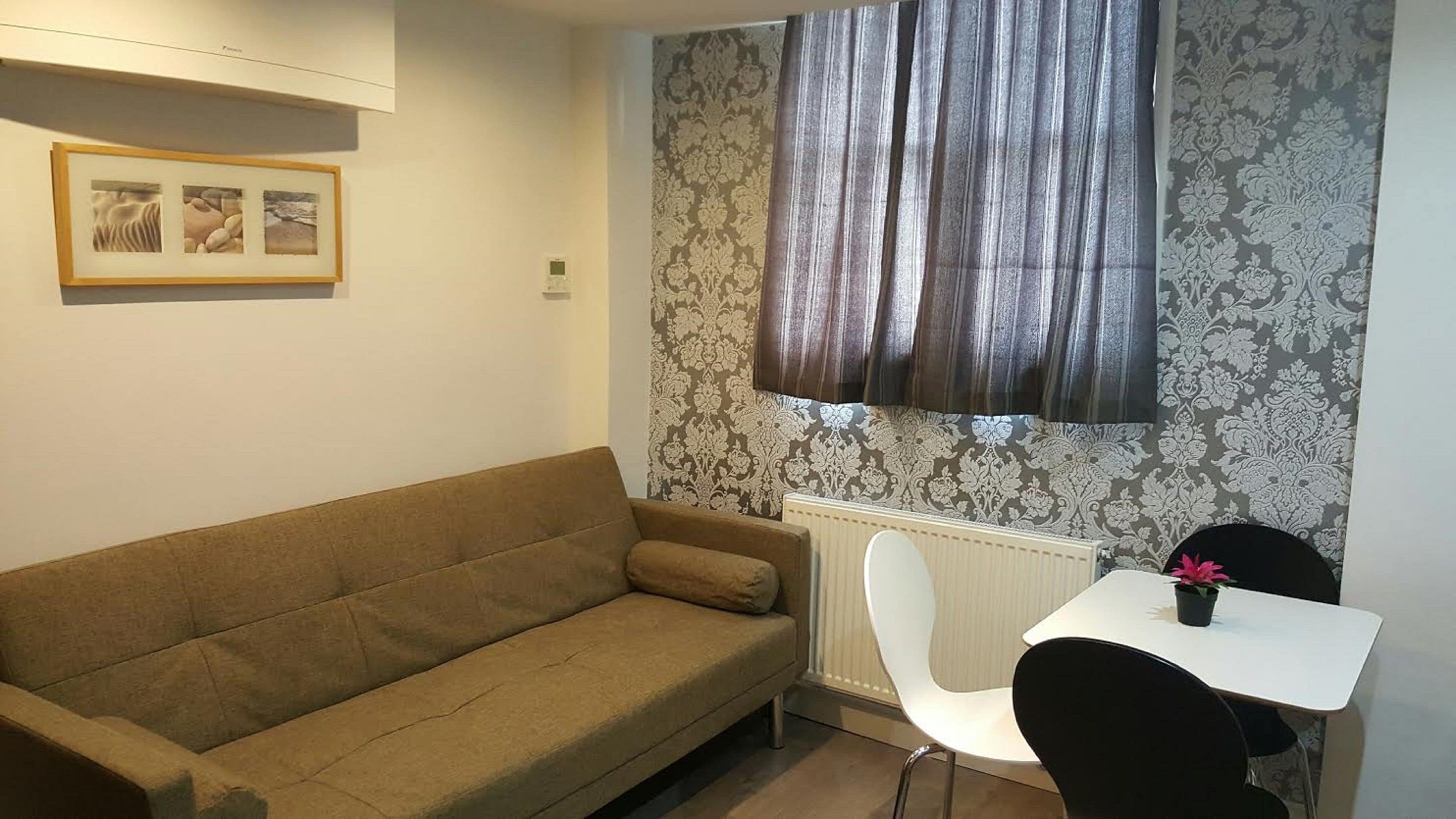 A room with a brown sofa, a white chair, a small table with a potted plant, and a window with patterned curtains.