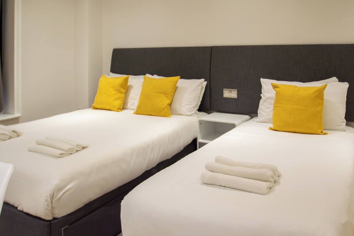 A hotel room with two beds, each with white linens and yellow pillows. There are folded towels on the beds.
