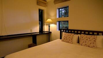 Una cama bien hecha con almohadillas florales, una mesita de noche con lámpara y una ventana con vistas a árboles.