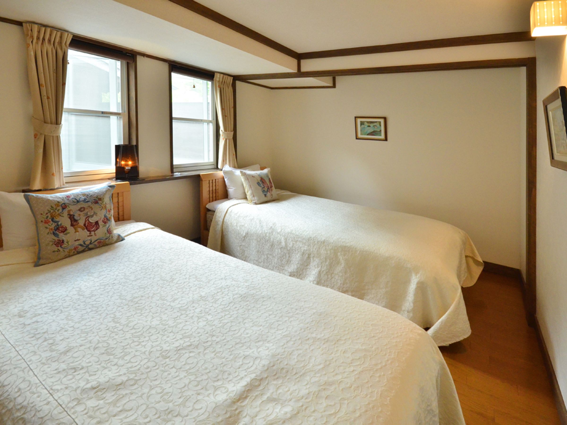 A room with two beds, a window with curtains, a small table with a lamp, and a framed picture on the wall.