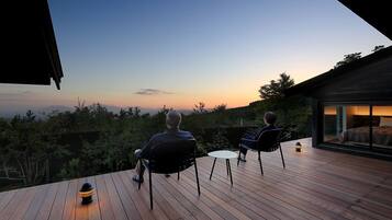 Terraza con dos sillas mirando una vista al atardecer, una pequeña mesa y un edificio moderno con paredes de vidrio.