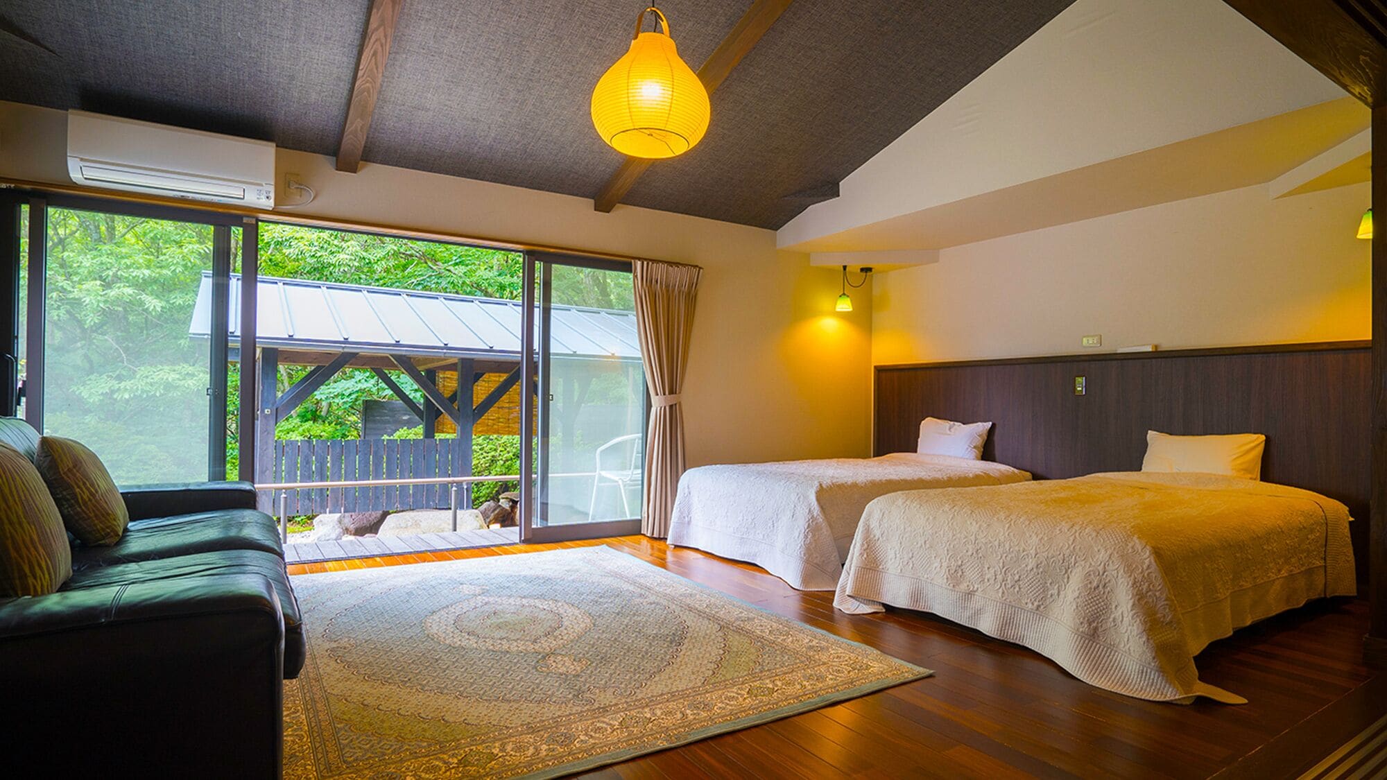 A room with two beds, a sofa, a large window with a view of greenery, and a hanging light fixture.