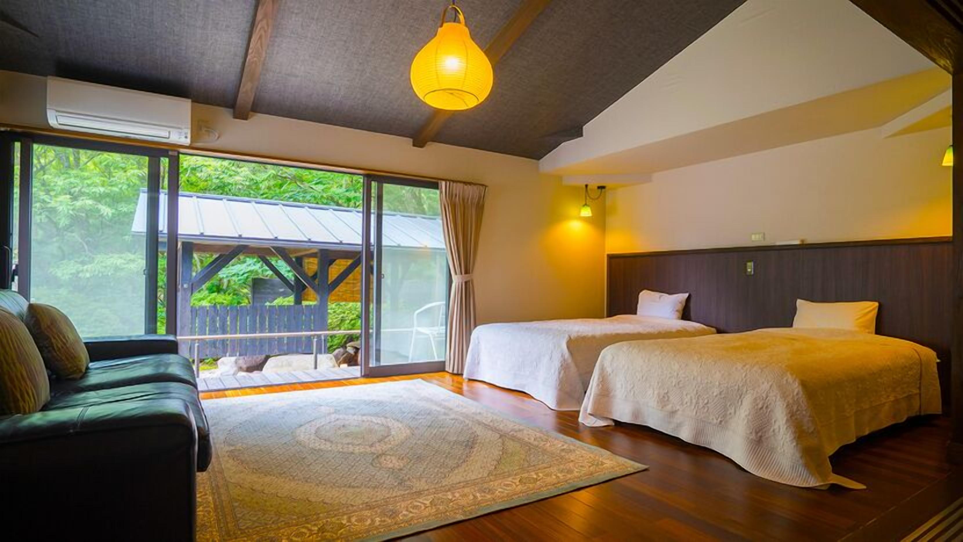 A room with two beds, a sofa, a large window with a view of greenery, and a hanging light fixture.