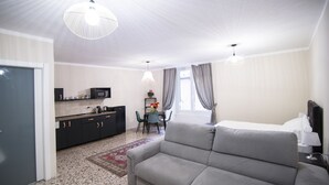 Una habitación de hotel moderna con sofá, una mesa de comedor con sillas, una zona de cocina y un gran ventanal con cortinas.