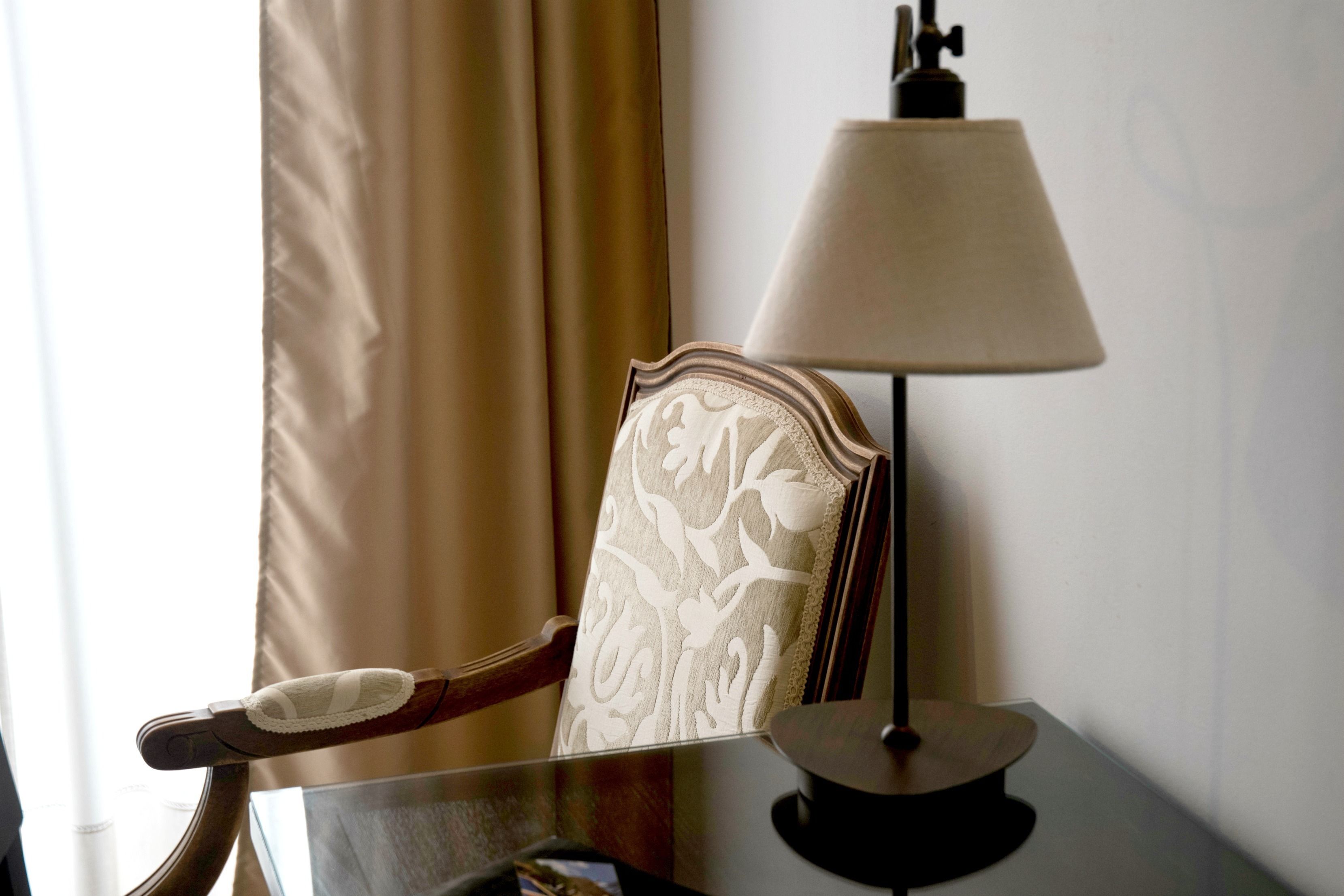 Une chaise avec un dossier en bois et un coussin, une table avec une lampe, et une fenêtre avec des rideaux.