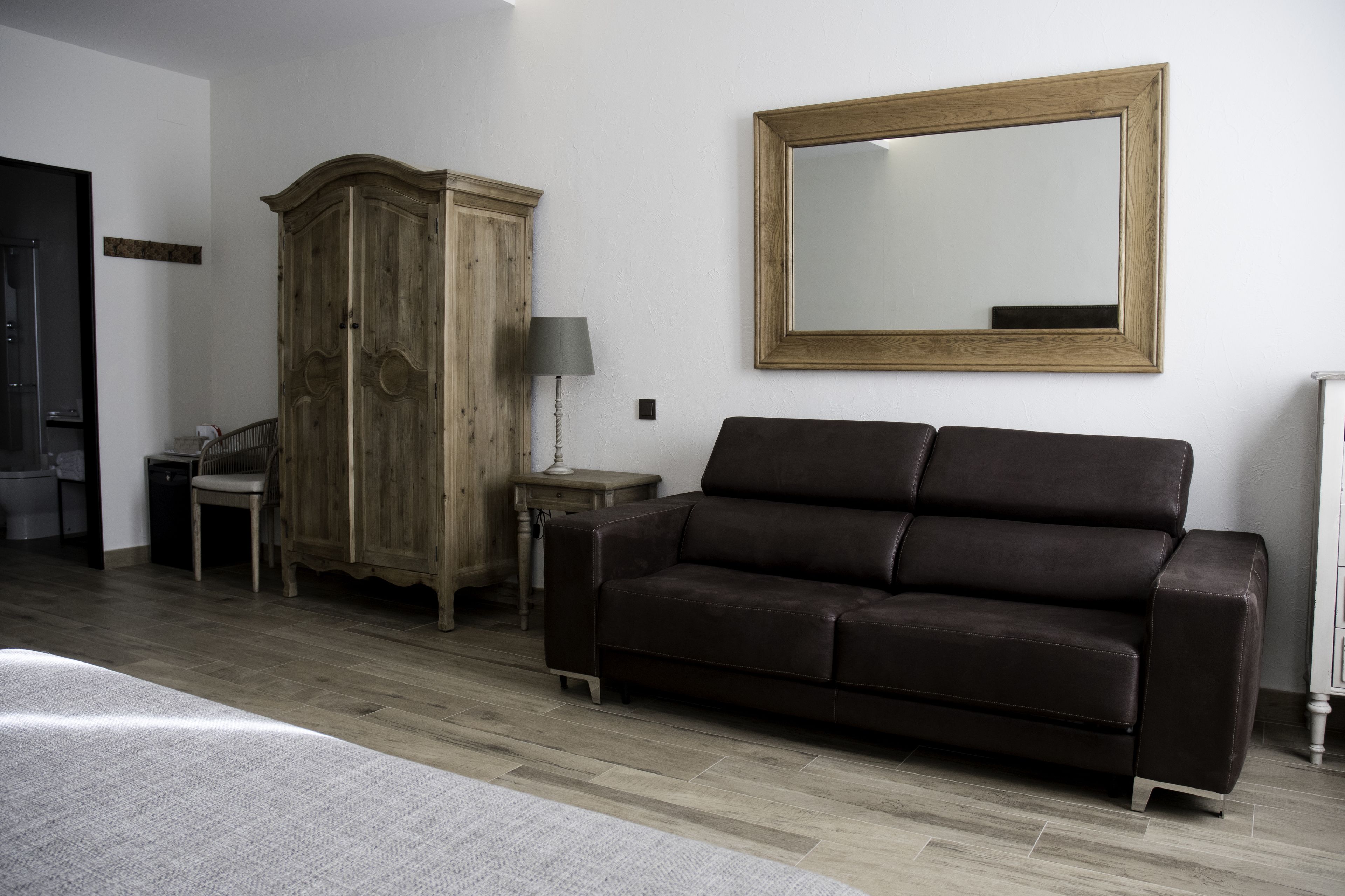 A bedroom with a wooden wardrobe, a dark brown sofa, a bedside table with a lamp, and a large mirror on the wall.