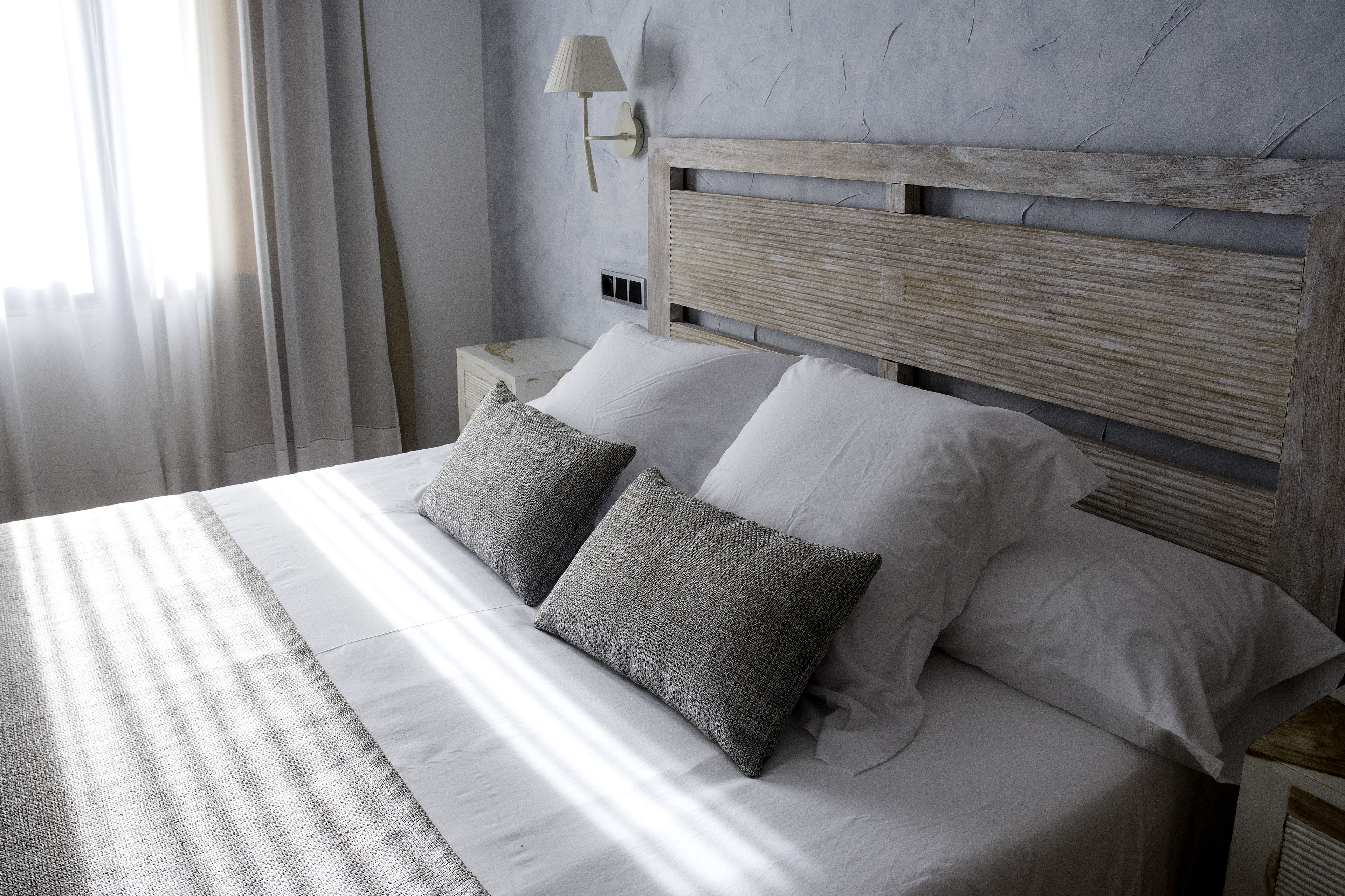 Una cama con cabecera de madera, dos almohadas blancas y una almohada gris con textura.