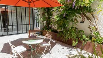 Un patio al aire libre con una mesa, sillas, un gran paraguas naranja y abundante vegetación.