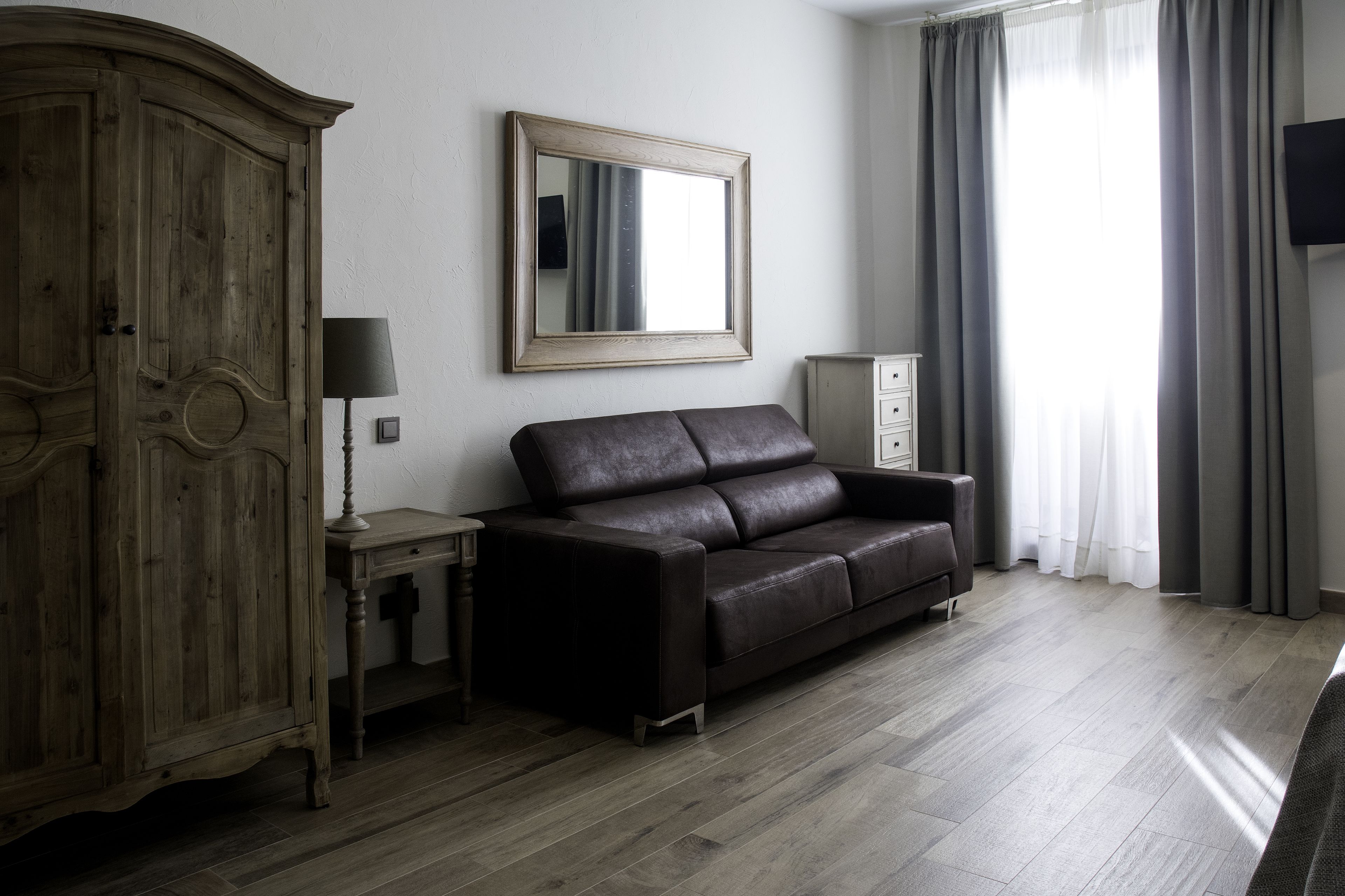 A living room with a wooden wardrobe, a dark leather sofa, a small table with a lamp, and a large mirror on the wall.