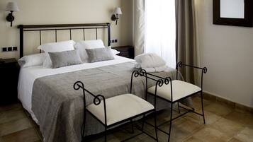 Una cama bien hecha con colcha gris y blanca, dos mesitas de noche con lámparas y una silla metálica con cojín.