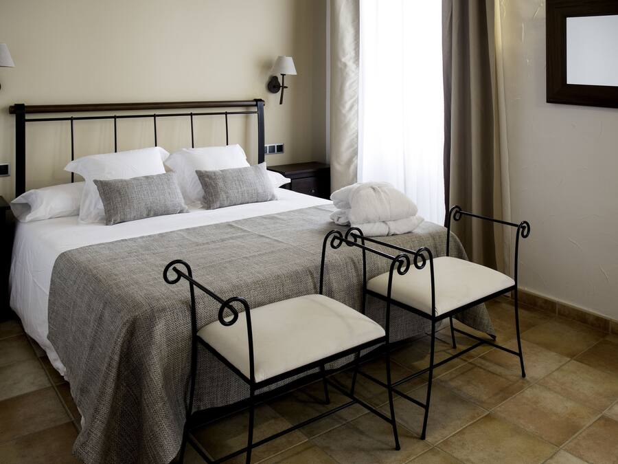 Una cama bien hecha con colcha gris y blanca, dos mesitas de noche con lámparas y una silla metálica con cojín.