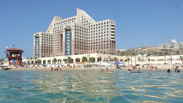 A large hotel with multiple floors and balconies, situated by a beach with clear blue water. - Apartment on the Beach (Haifa)