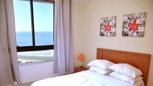 A hotel room with a large bed, a nightstand, and a window with a view of the beach. - Apartment on the Beach (Haifa)