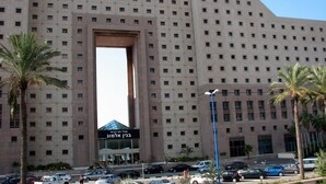 A large building with a distinctive arch entrance, multiple floors, and a parking area with cars and palm trees. - Apartment on the Beach (Haifa)
