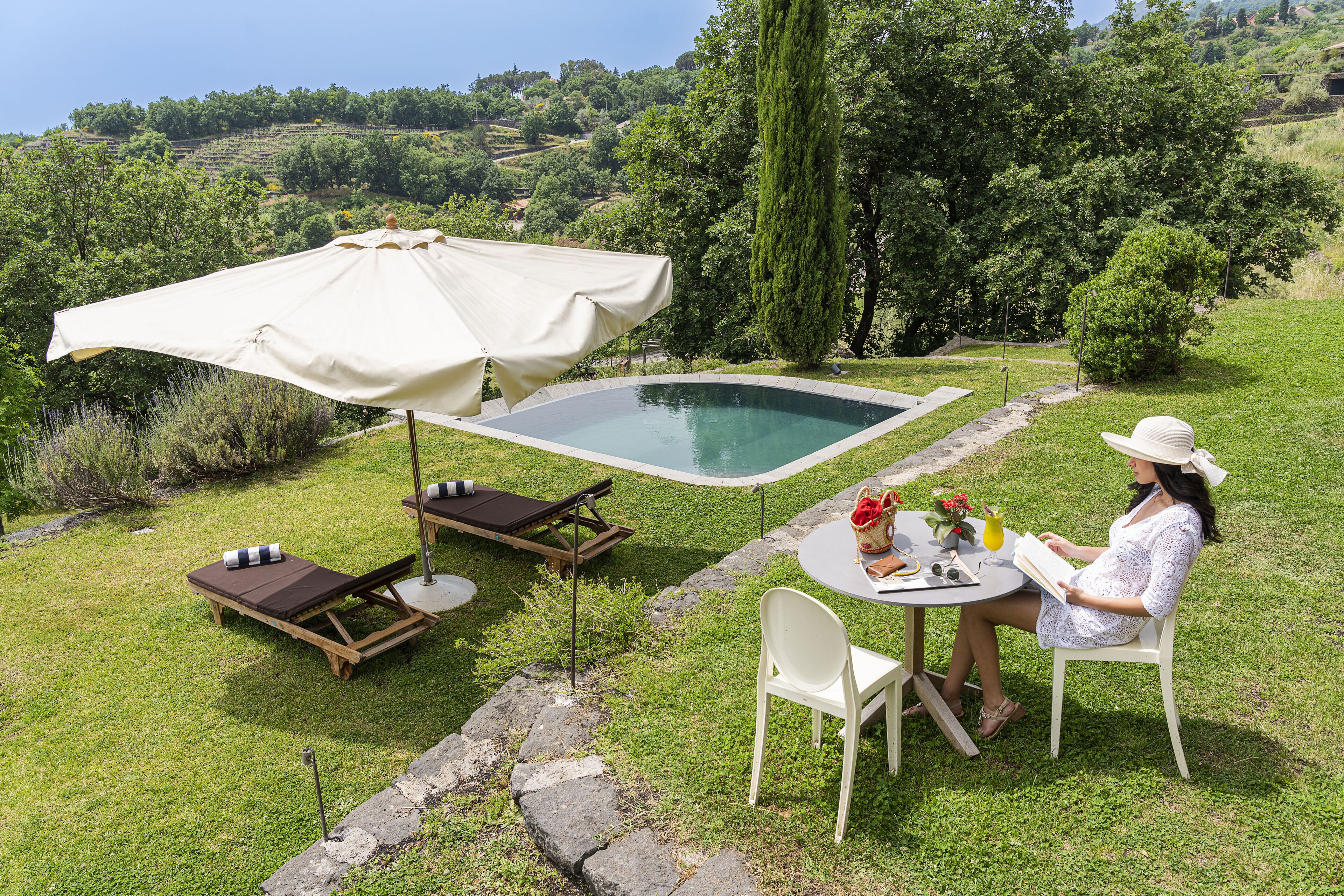 A poolside area with a lounge chair, a table with a drink, and a person reading a book.