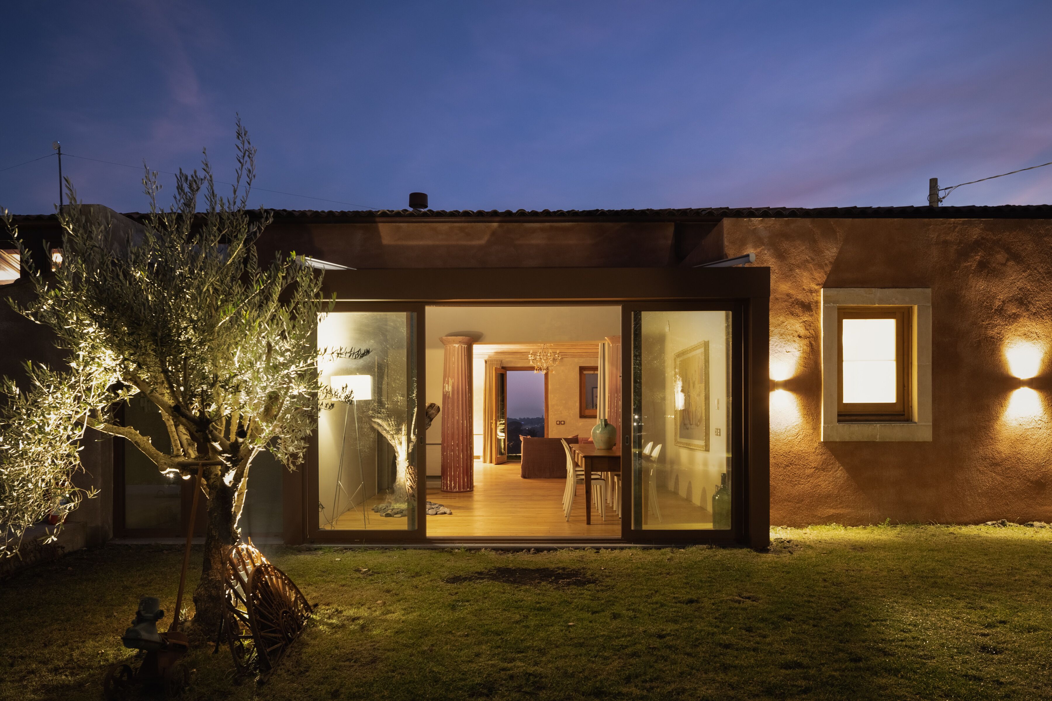 A modern house with a glass door entrance, a dining area with a table and chairs, and a large tree in the yard.