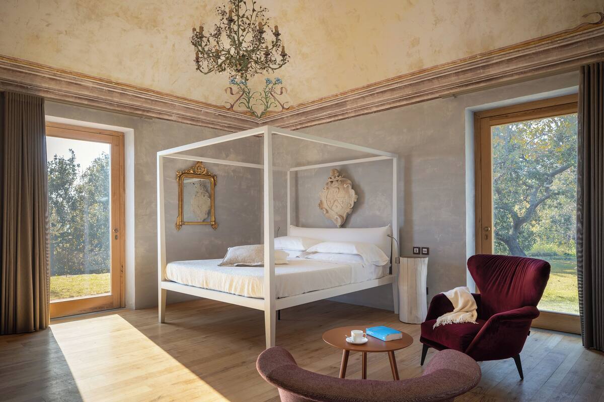 Une chambre avec un lit à baldaquin, un fauteuil rouge, une petite table et une grande fenêtre donnant sur des arbres.