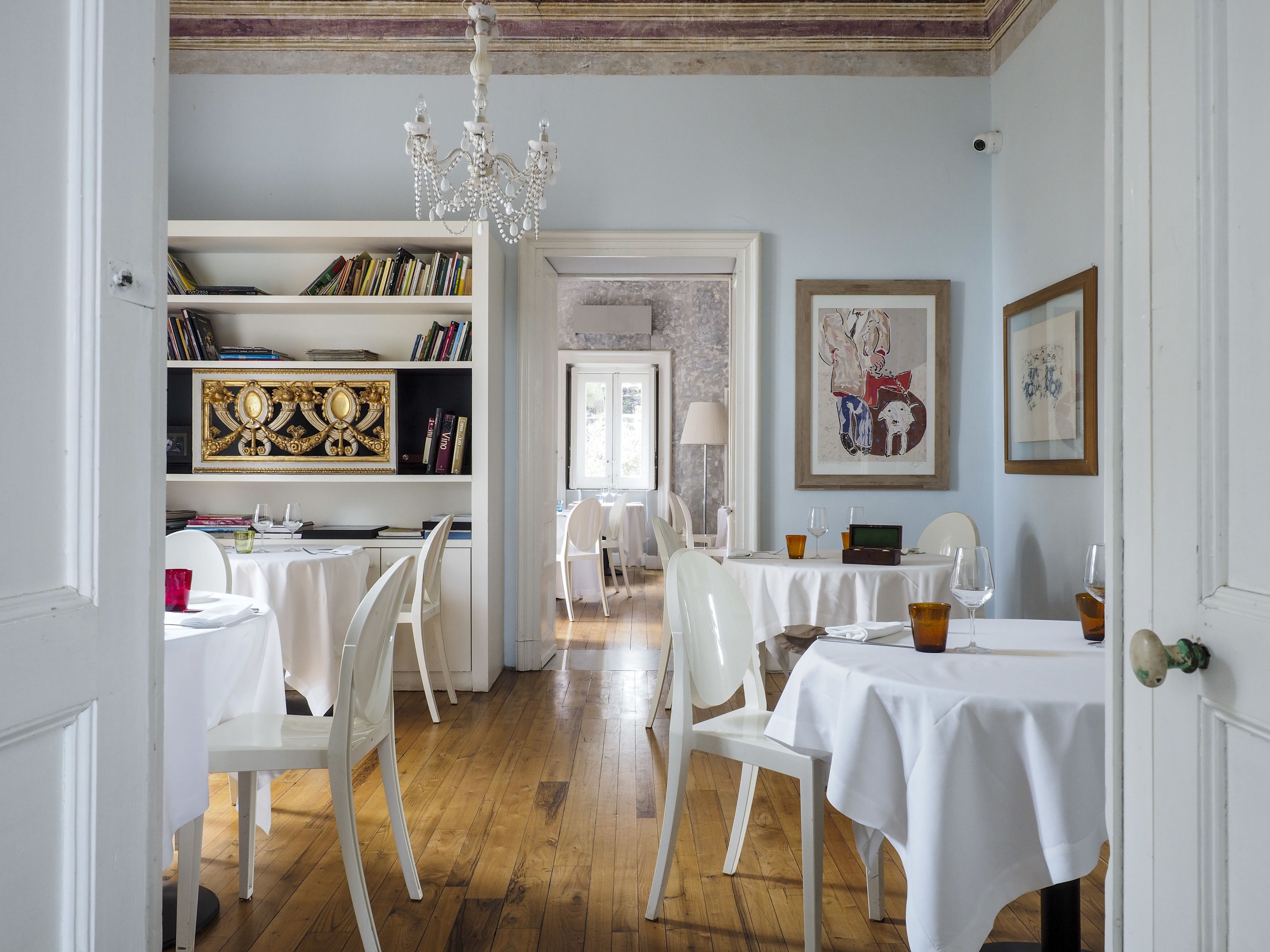 A dining area with white tables and chairs, a chandelier, and a bookshelf with books and decorative items.