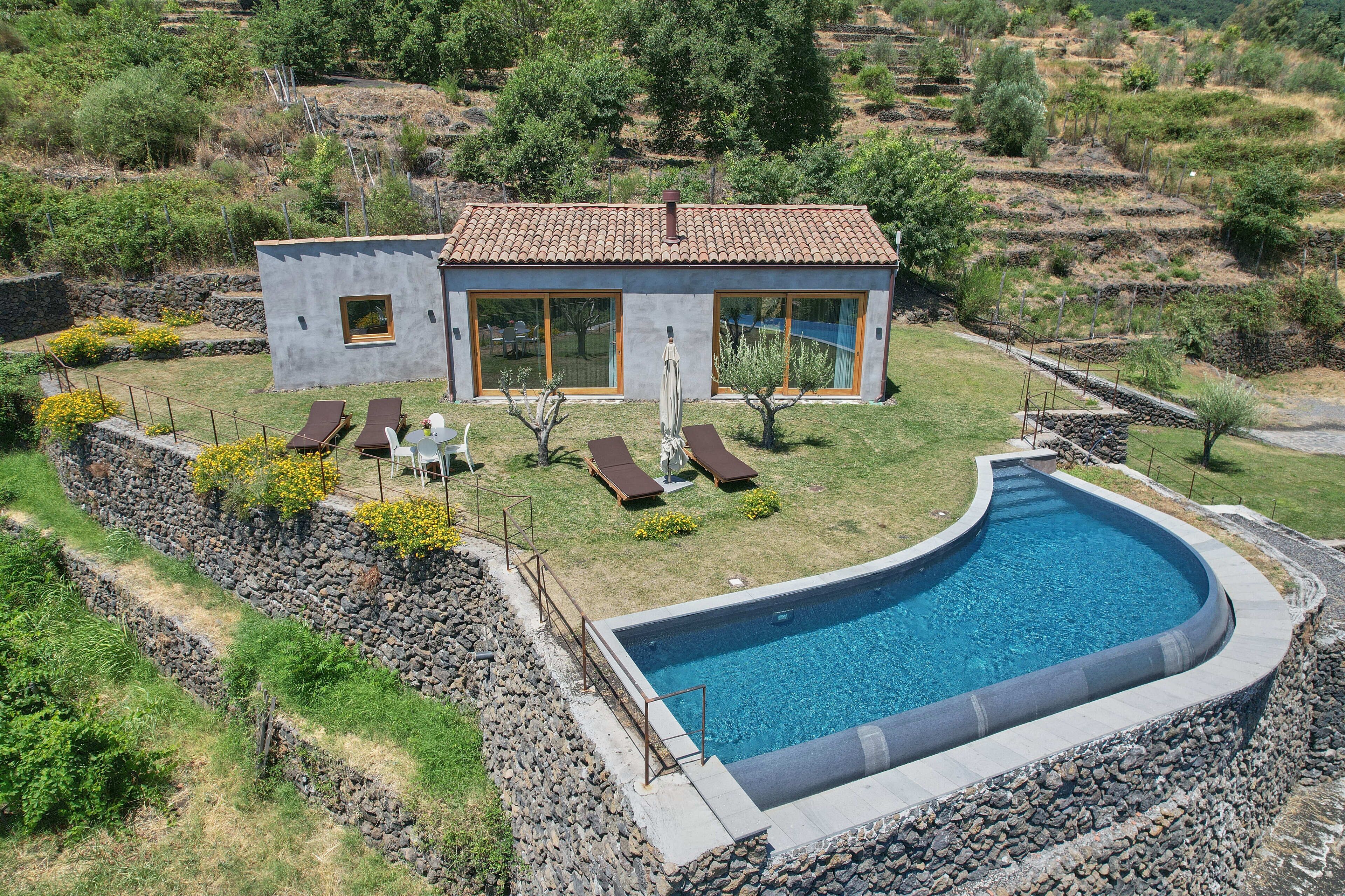 A modern house with a swimming pool, surrounded by a stone wall and a grassy area with sun loungers.
