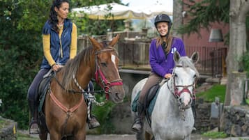Due persone a cavallo in un ambiente esterno, con vegetazione e un muro di pietra sullo sfondo.