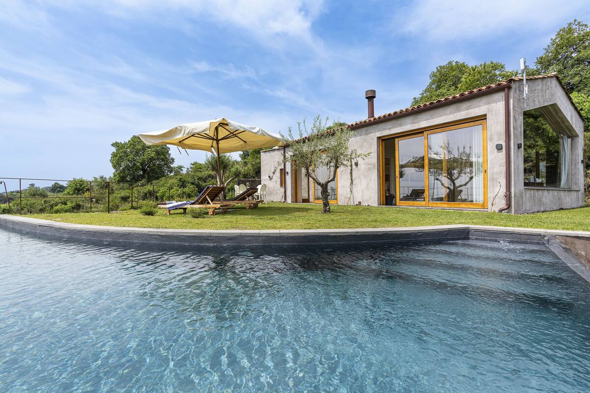 Une piscine avec un transat et un parasol, un petit bâtiment doté de grandes fenêtres, et un arbre en arrière-plan.