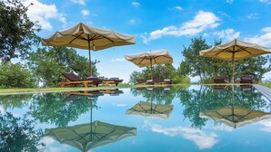 Zona de piscina con sillones de descanso y sombrillas, rodeada de árboles.