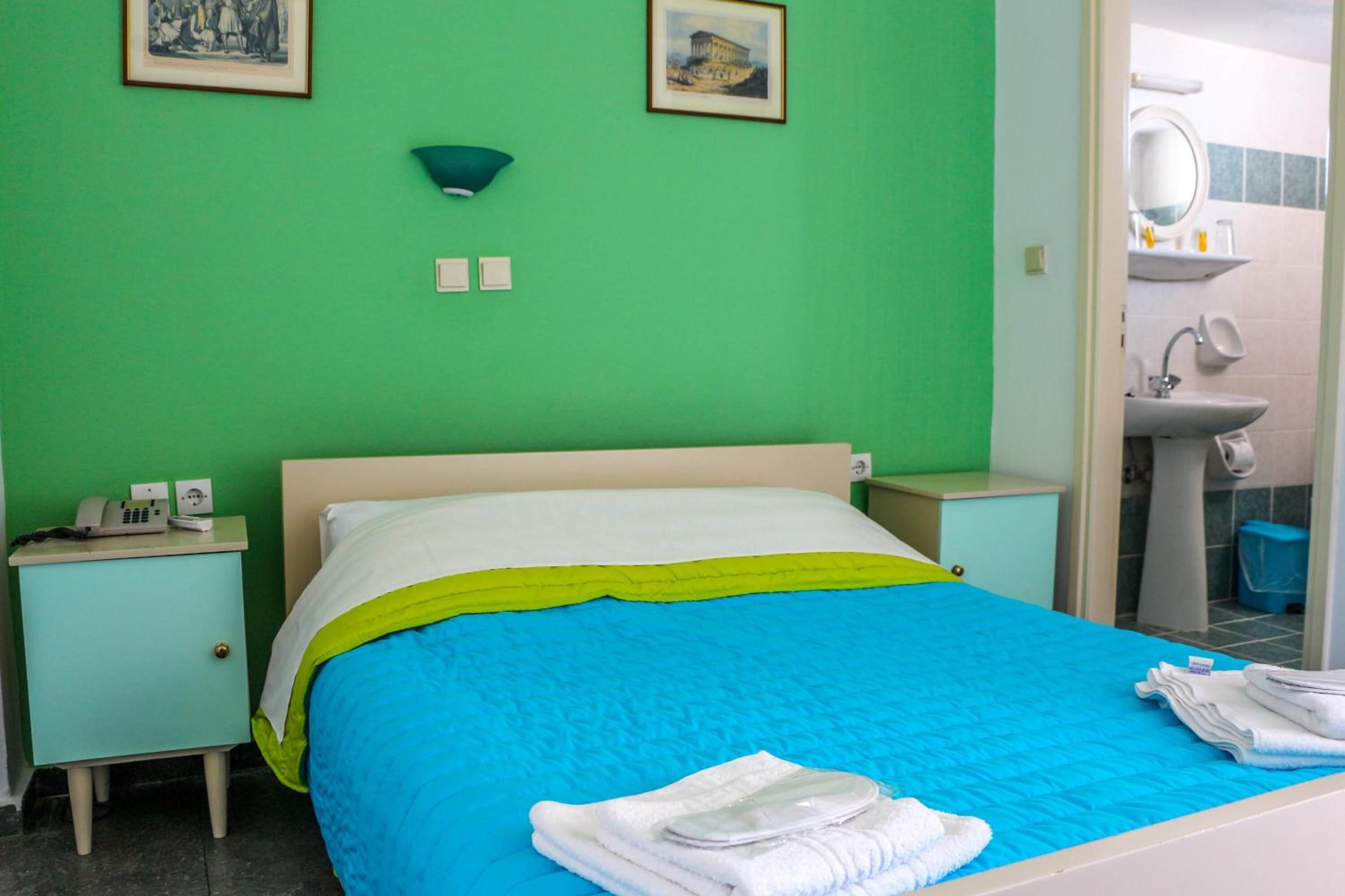 A hotel room with a bed, a nightstand, a bedside table, a mirror, a sink, and a towel rack.