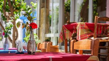 A table set outdoors with a red tablecloth, wooden chairs, and a vase with red flowers.