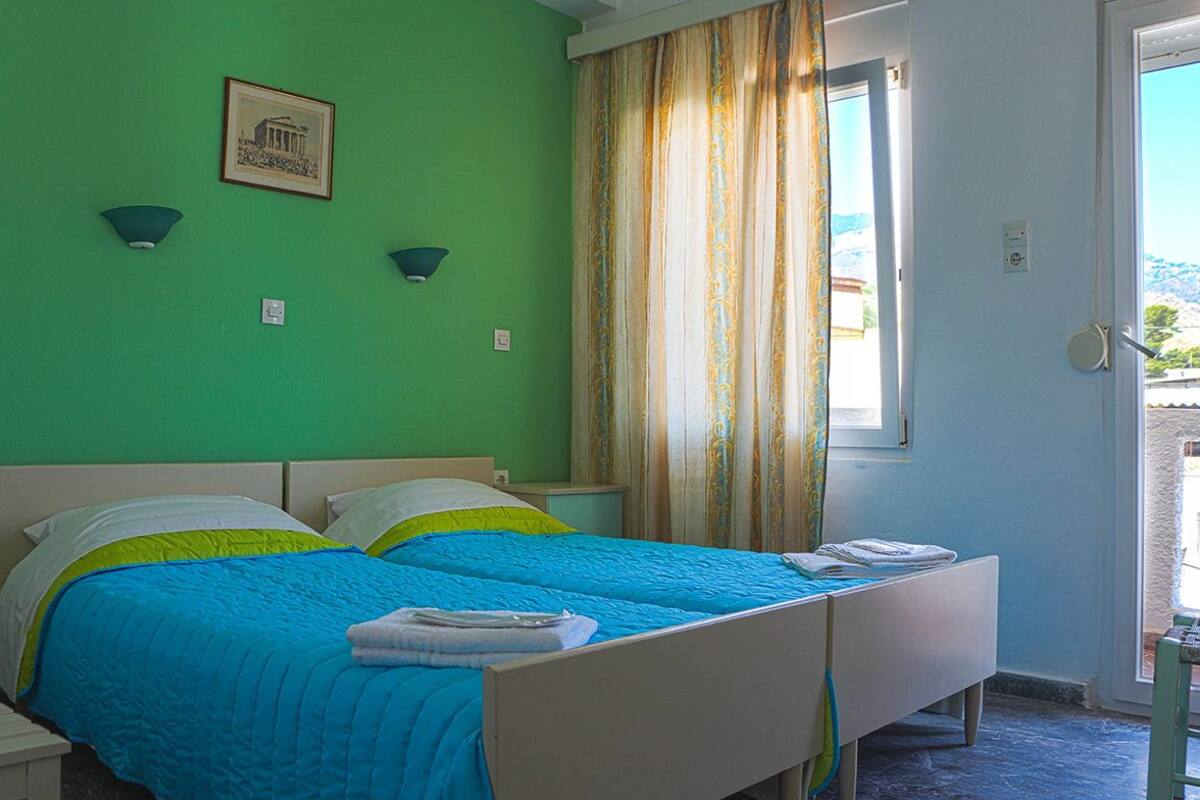 A double bed with blue and green bedding, a small bedside table, and a window with curtains.