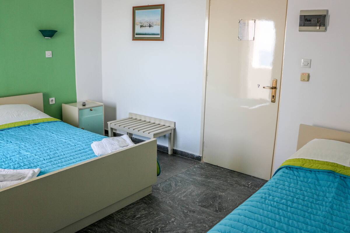 A room with two beds, a small table, a nightstand, a door, and a wall-mounted light fixture.
