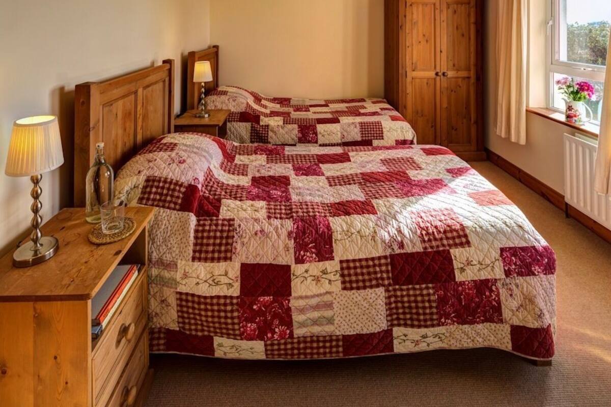 Ein Schlafzimmer mit zwei Betten, Holz-Kopfteilen, einem Nachttisch mit Lampe, einer hölzernen Kommode und einem Fenster mit Blick.