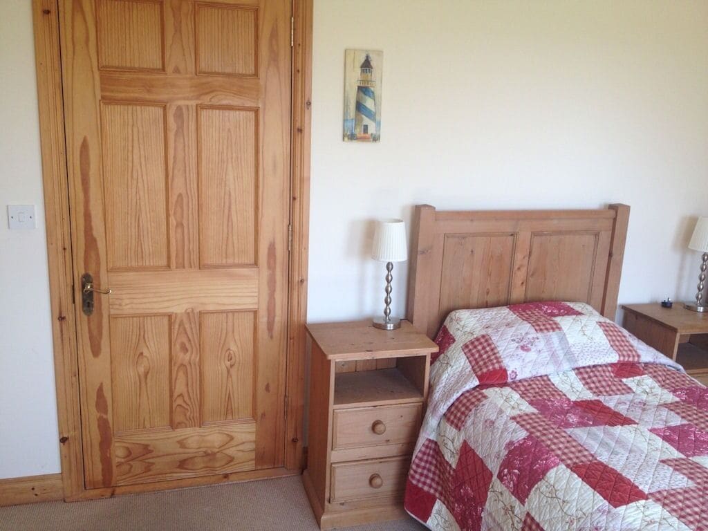 Une chambre avec une porte en bois, un lit recouvert d’une courtepointe à carreaux, une table de chevet avec une lampe et un petit tableau accroché au mur.