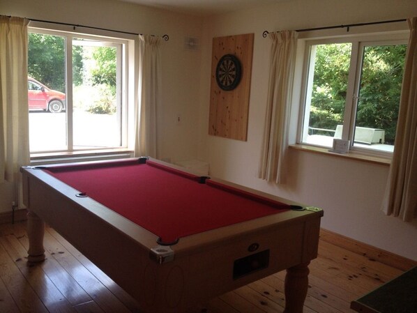 Una mesa de billar en una habitación con suelo de madera, dos ventanas con cortinas y un dardo en la pared.