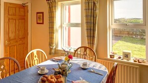 Una mesa de comedor con mantel azul, una bandeja de pasteles, un calentador de agua y una taza. Hay sillas alrededor de la mesa, y una ventana con cortinas.