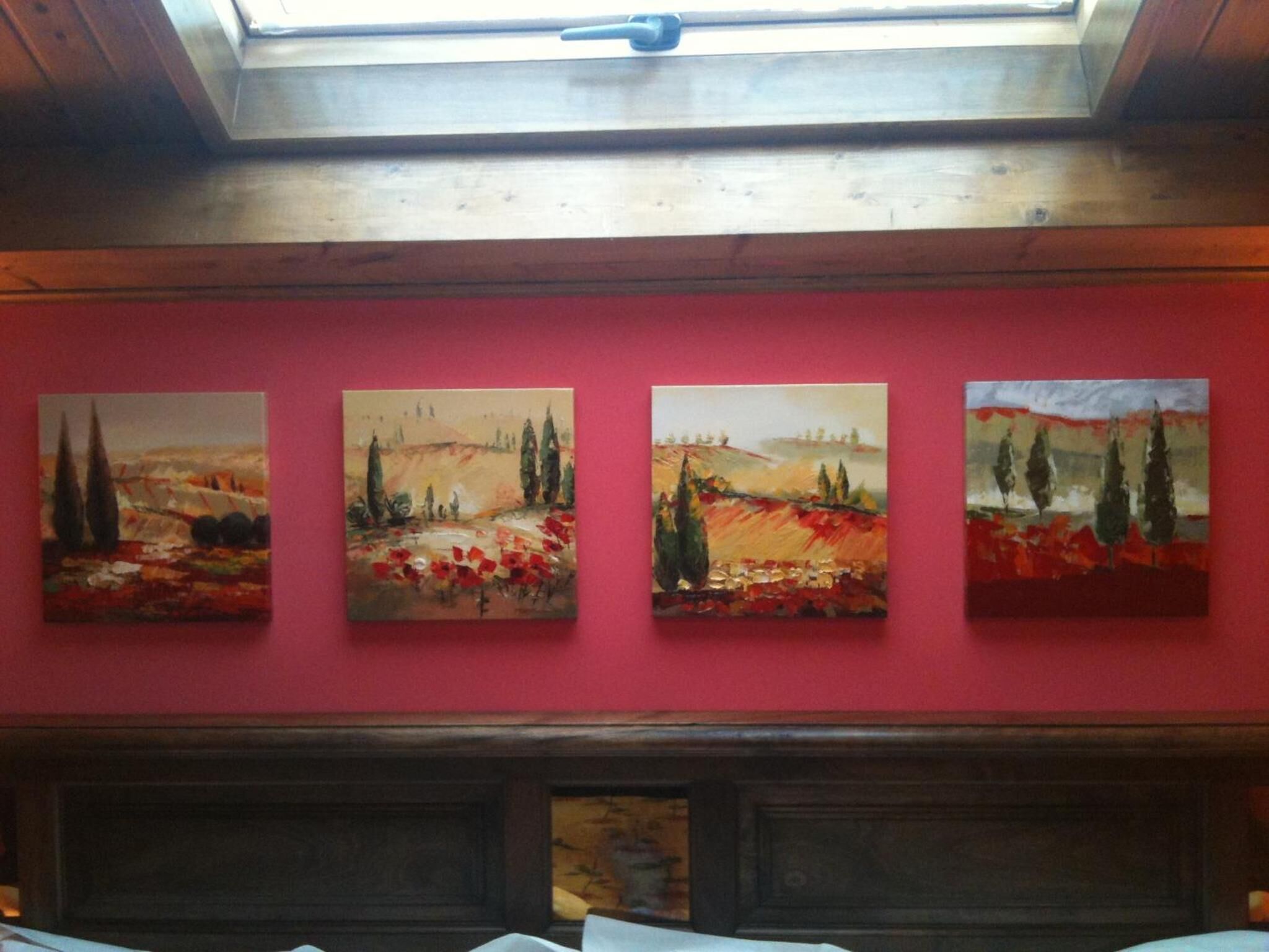 Una habitación con tragaluces, una pared roja y cuatro cuadros que muestran paisajes.