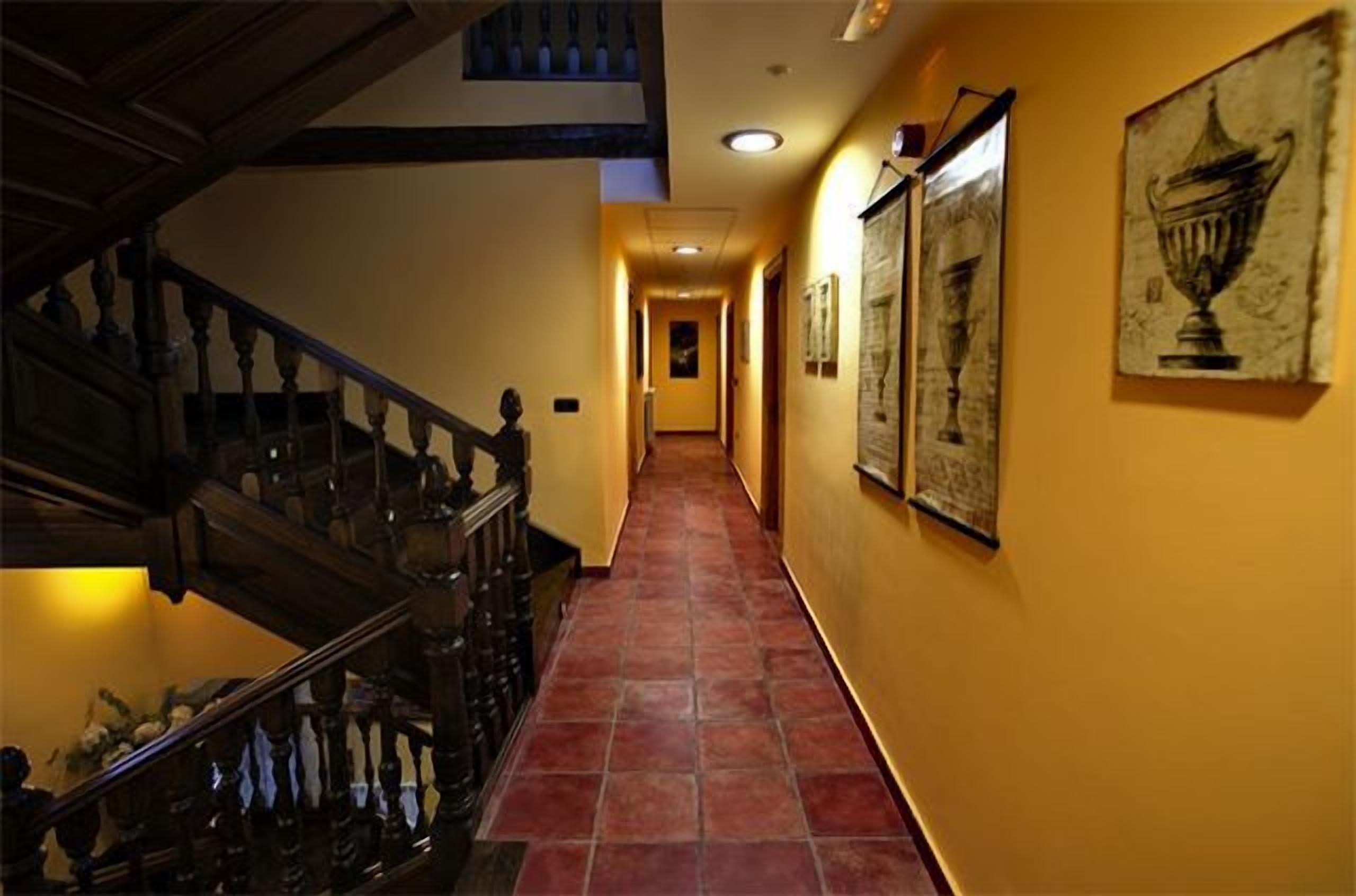 Un pasillo con una escalera de madera a la izquierda, suelo de baldosas y cuadros en las paredes.