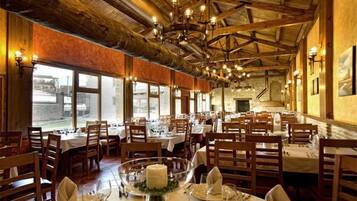 Un restaurante con vigas de madera y candelabros, mesas dispuestas con manteles blancos y velas, y ventanas amplias con vistas.