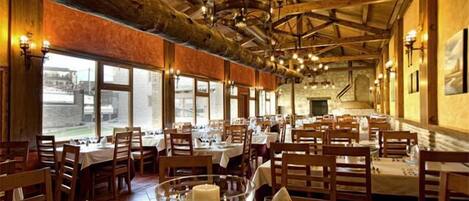 A restaurant with wooden beams and chandeliers, tables set with white tablecloths and candles, and large windows offering a view.