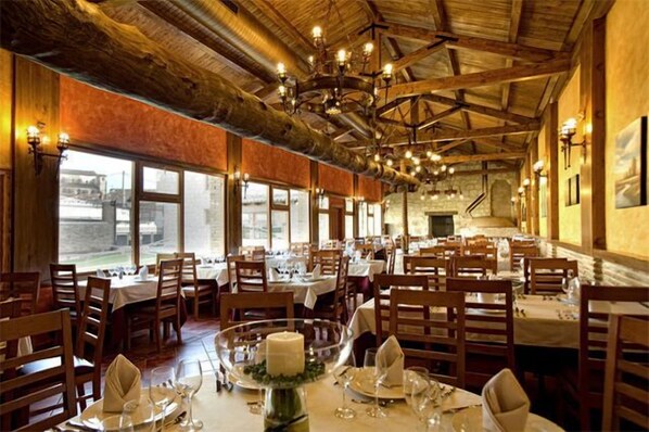 A restaurant with wooden beams and chandeliers, tables set with white tablecloths and candles, and large windows offering a view.