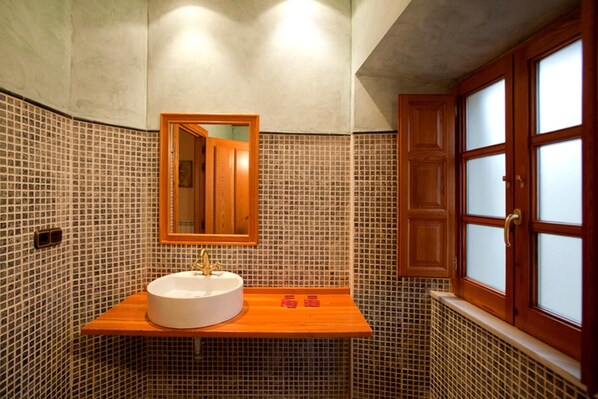 Un baño con pared de azulejos, tocador de madera, lavabo redondo, espejo y un mueble con puertas de vidrio esmerilado.