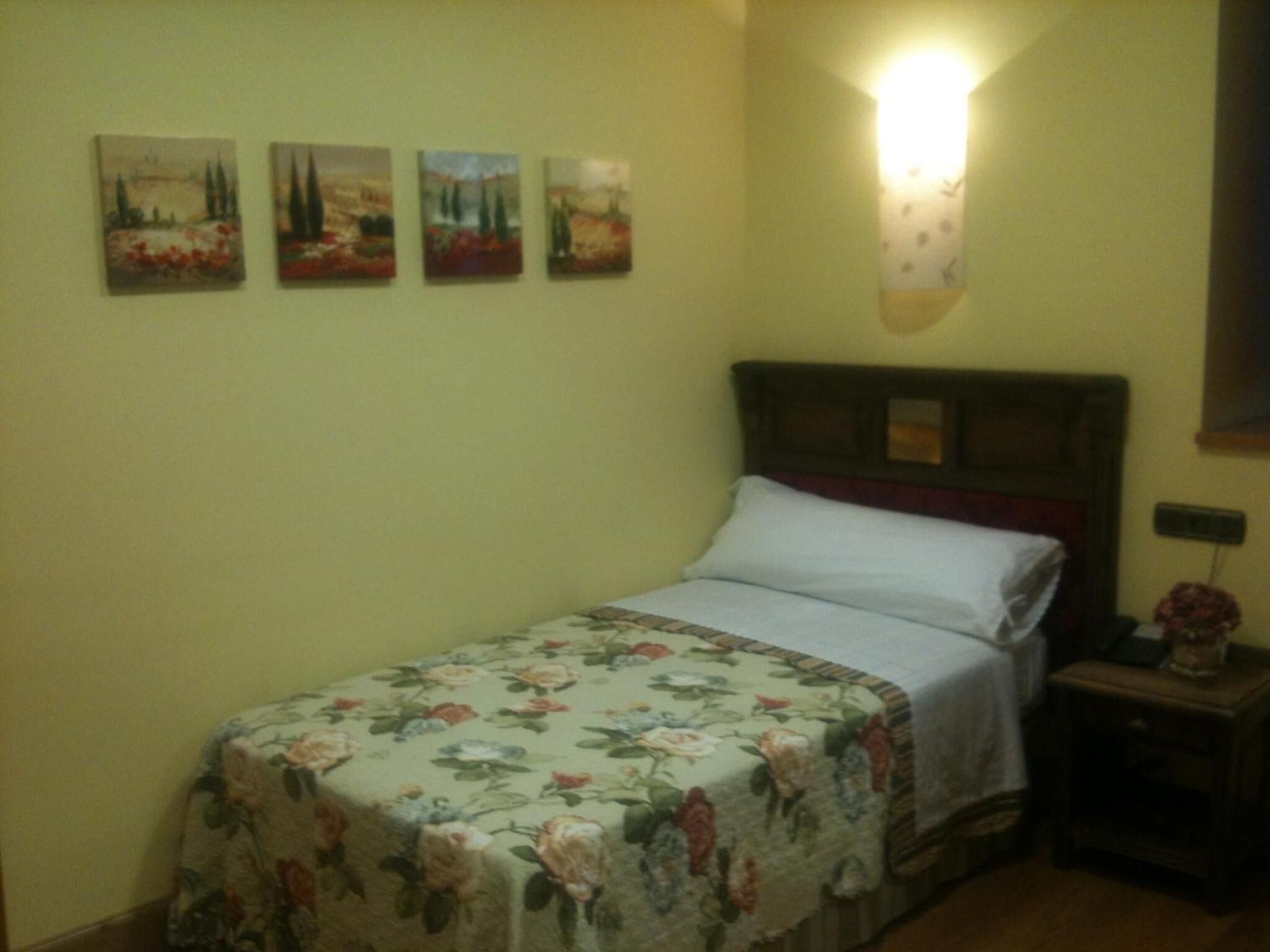 Un dormitorio con cama, colcha estampada, una mesita de noche con lámpara y cuatro cuadros de paisajes en la pared.