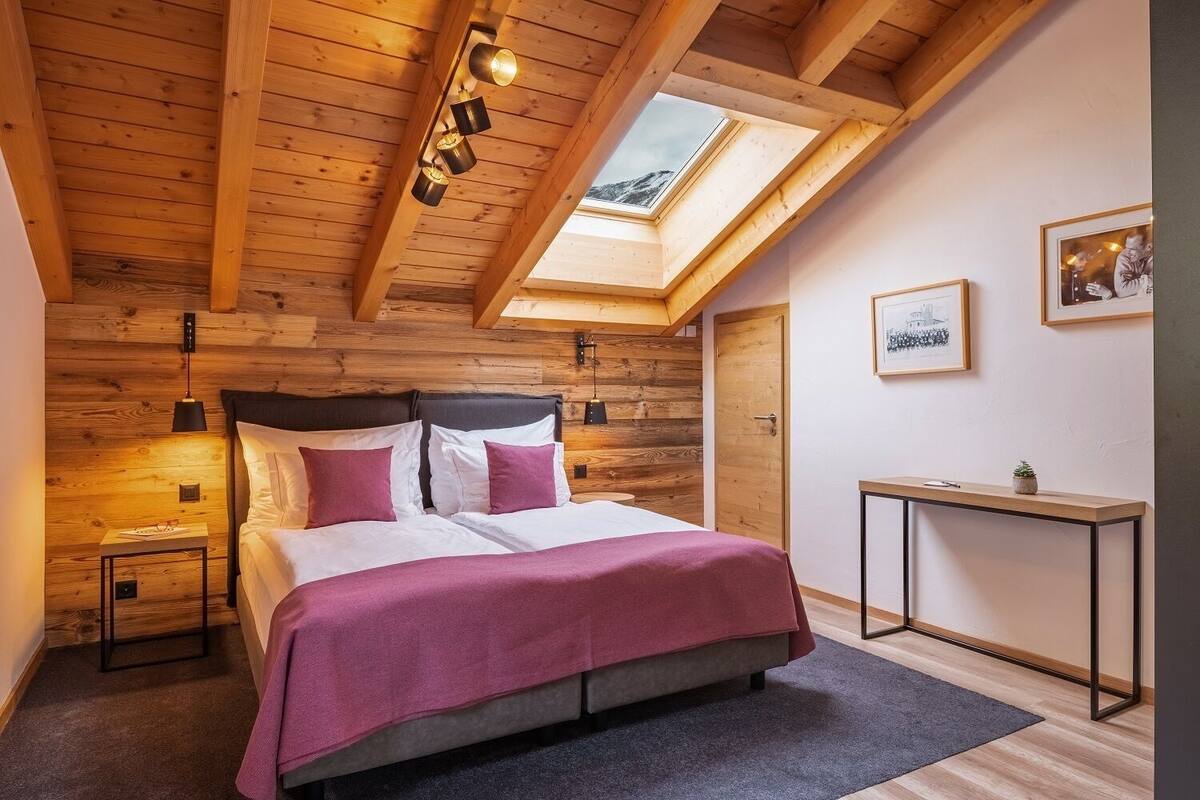 Ein Schlafzimmer mit Holzbalkendecke, einem Oberlicht, einem Bett mit weißer und violetter Bettwäsche, einem Nachttisch und einem kleinen Schreibtisch mit Pflanze.