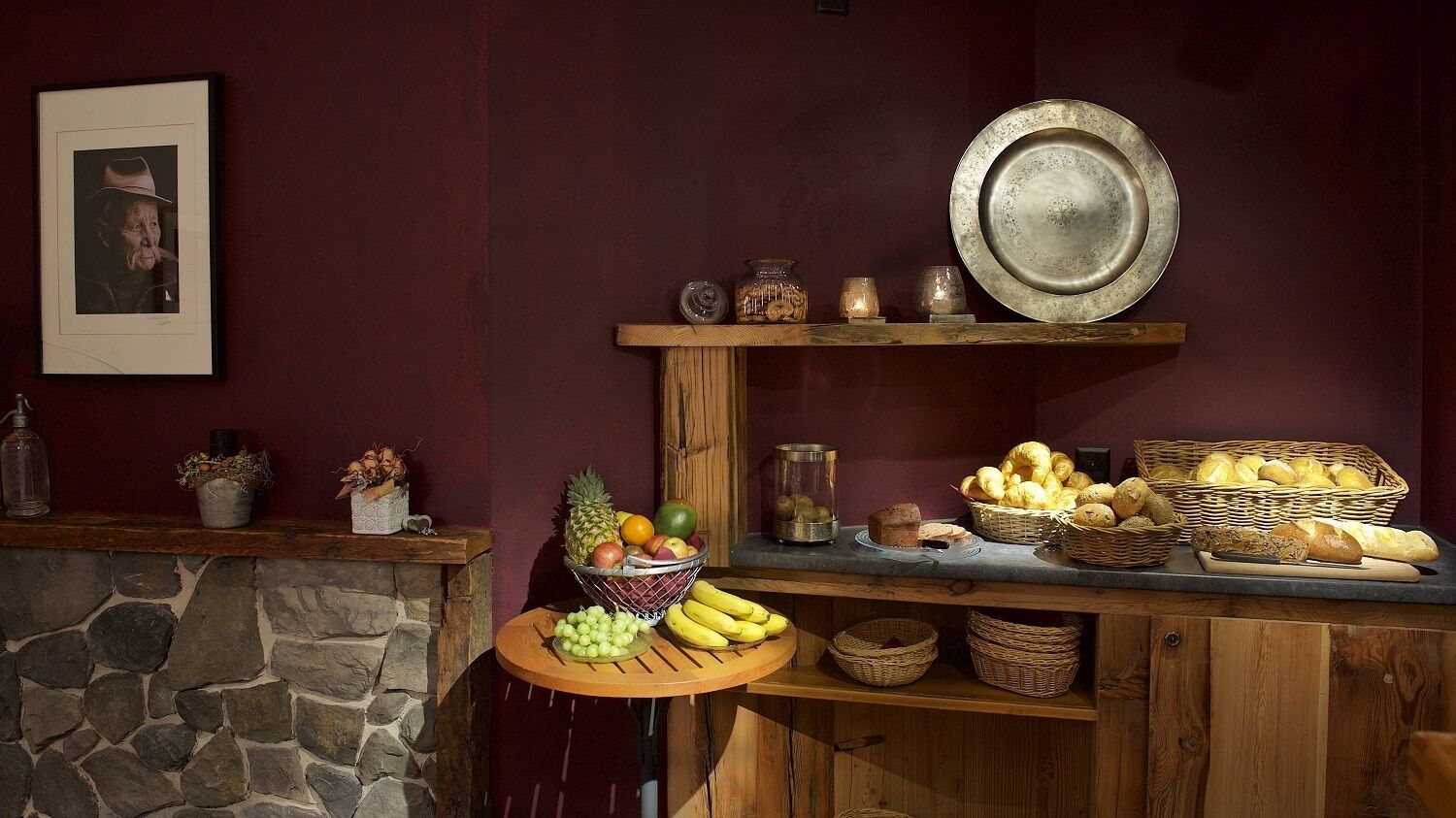 Une cuisine rustique aux murs de pierre, aux étagères en bois et où l’on trouve une variété de fruits et de pains.