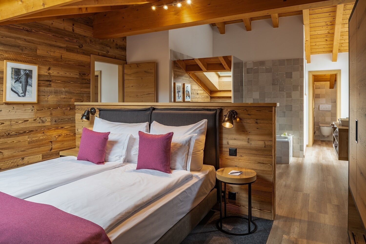 Un dormitorio con cabecera de madera, dos camas, una mesita redonda y un baño visible a través de una puerta abierta.