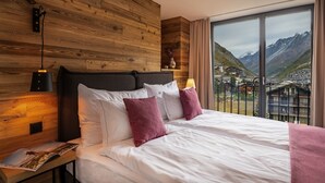 Une chambre à coucher avec une tête de lit en bois, un grand lit avec des oreillers blancs et roses, une table de chevet avec une lampe, et une vue sur les montagnes par une grande fenêtre.